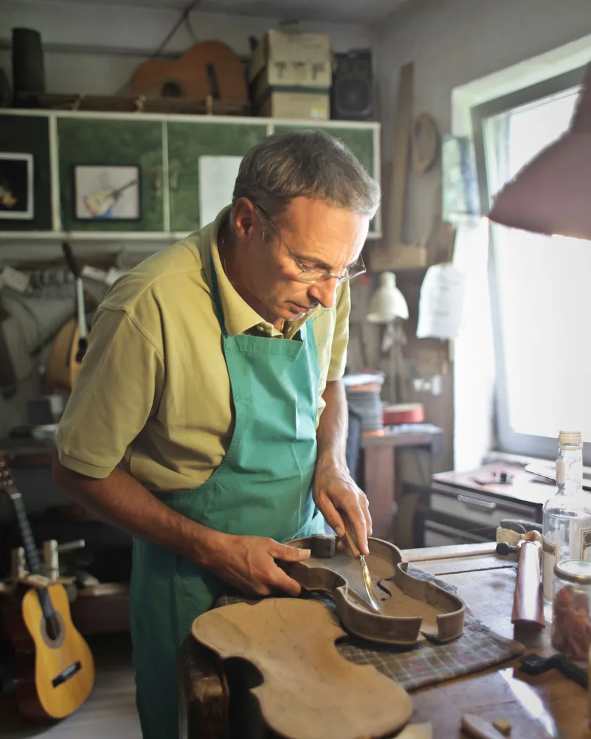 Manos trabajando una pieza de madera de violín sujeta en el banco, con herramientas de corte en primer plano